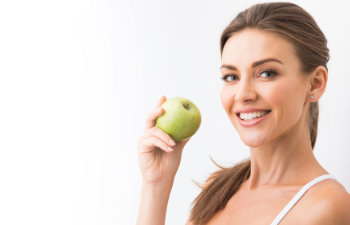 A person with light brown hair smiles at the camera and holds a green apple.