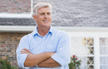 A man with gray hair, wearing a light blue shirt, stands with arms crossed in front of a brick house with white-framed windows.