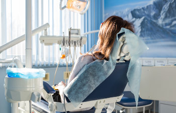 A woman with brown hair sits in a dental chair covered with a blue disposable sheet, facing away from the camera in a brightly lit dental office with mountain scenery on the wall.